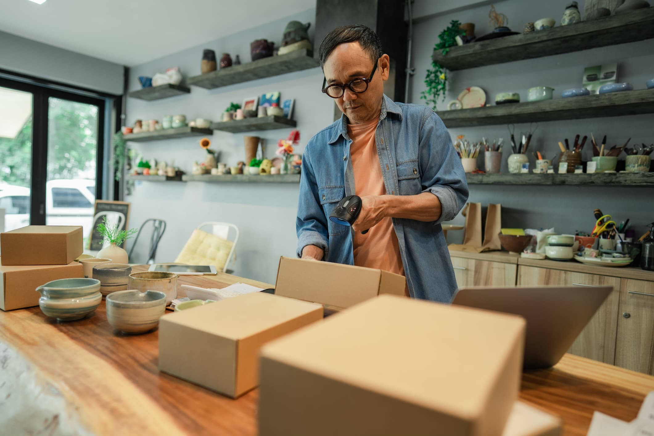 A potter in his studio scanning the barcode on a shipping box, his ceramics on the shelves behind him