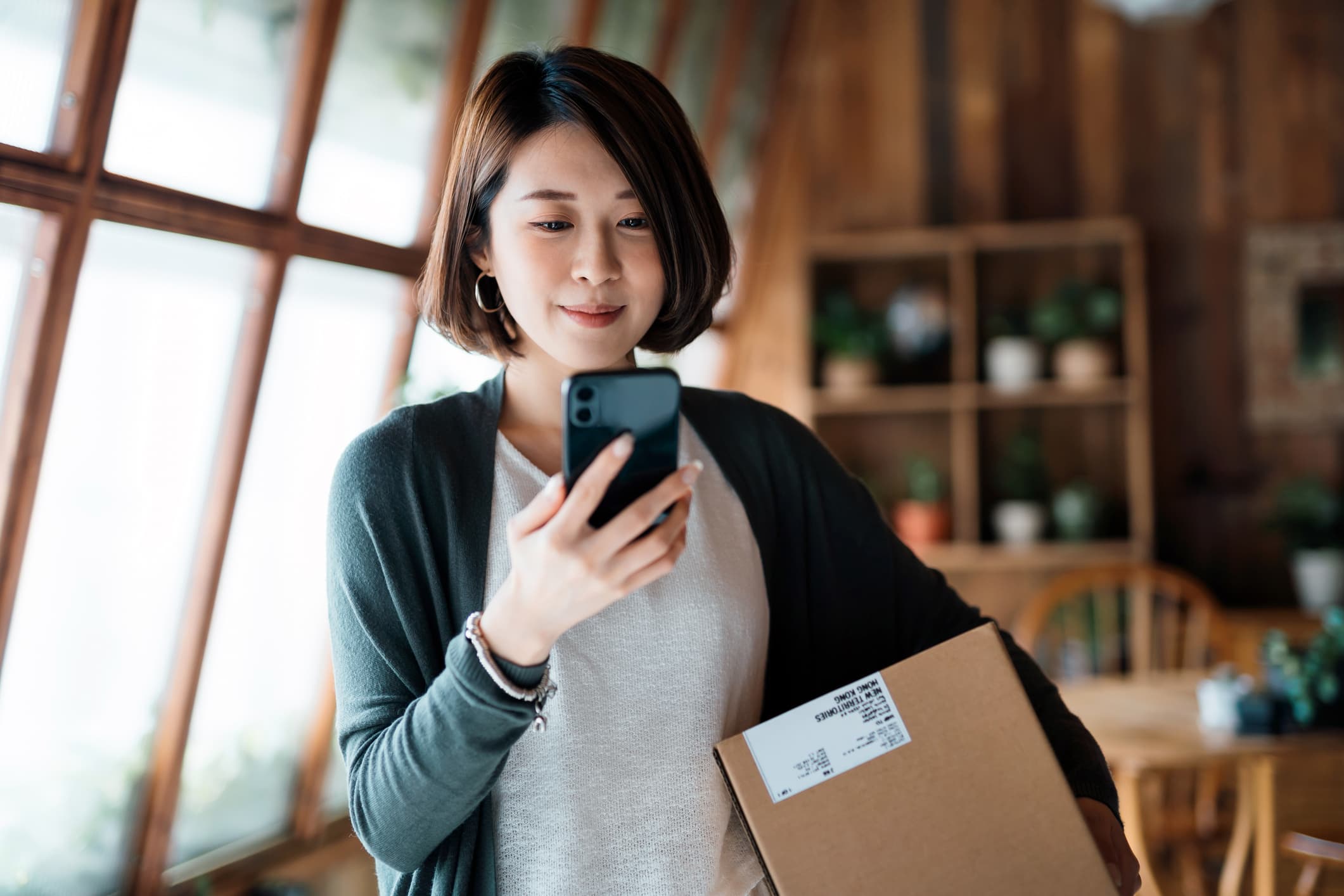 A woman smiling at her phone with a delivered parcel under her arm, standing by a sunny window at home.